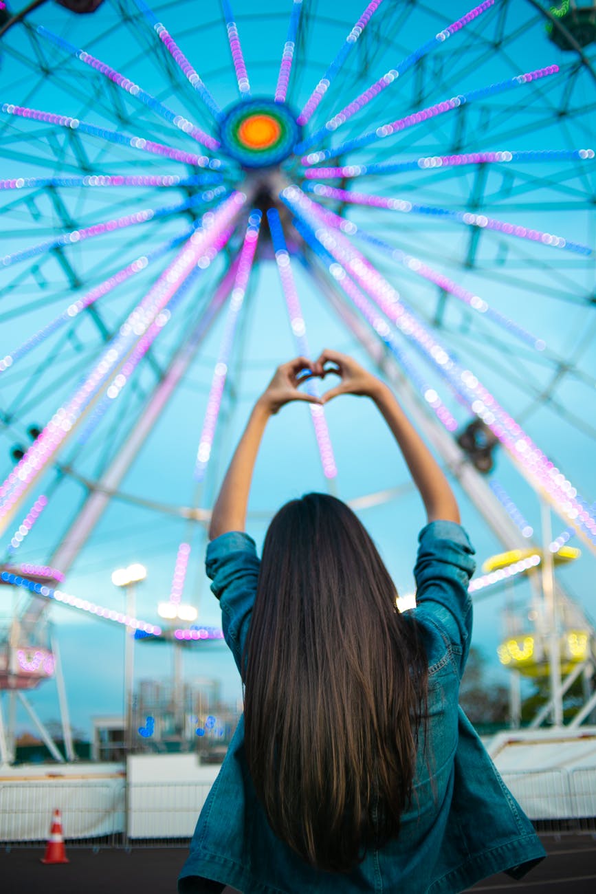 woman facing ferris wheel while making heart hand sign